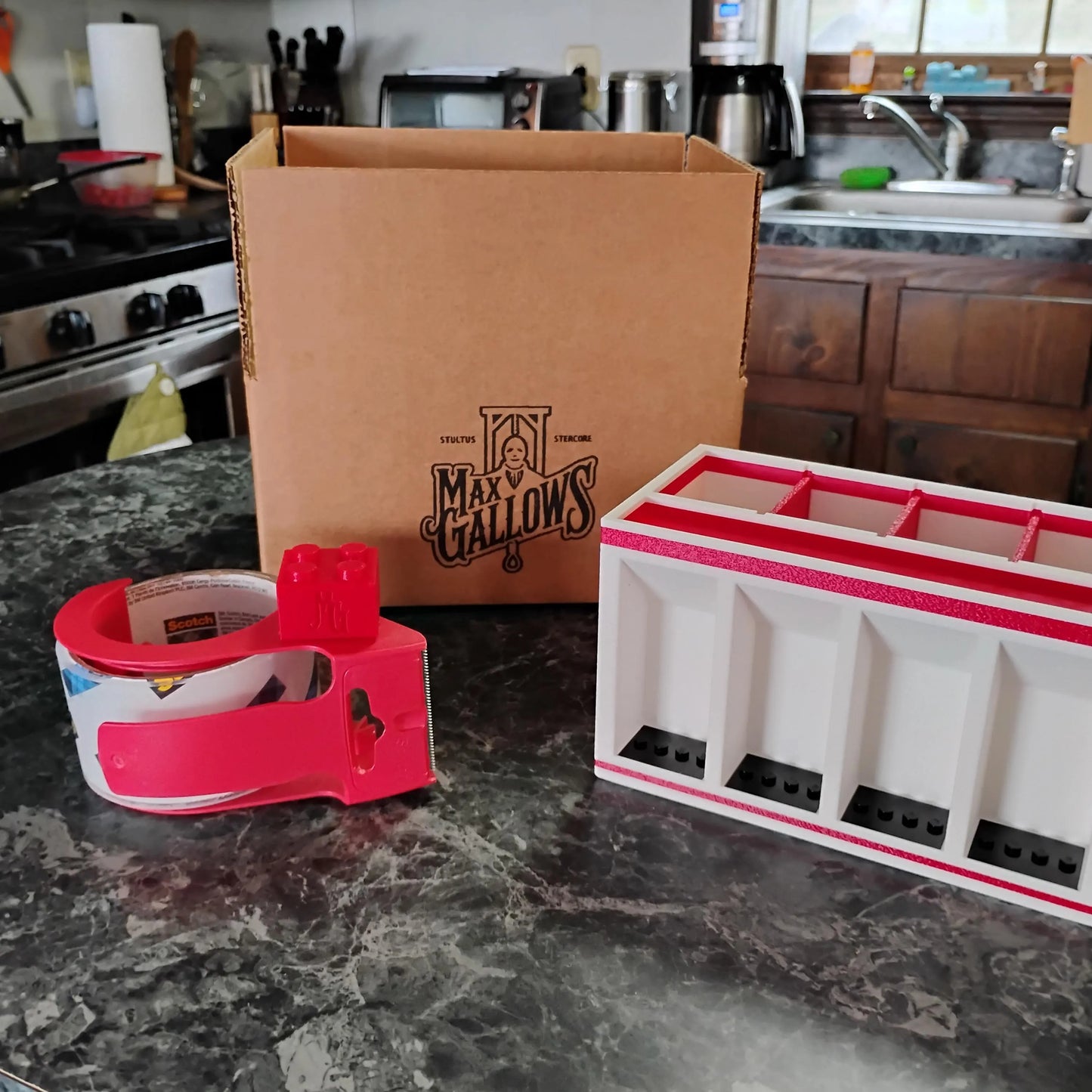 Kitchen counter with Max Gallows product, tape dispenser, and white Desk Organizer/Minifigure Display with red accents.