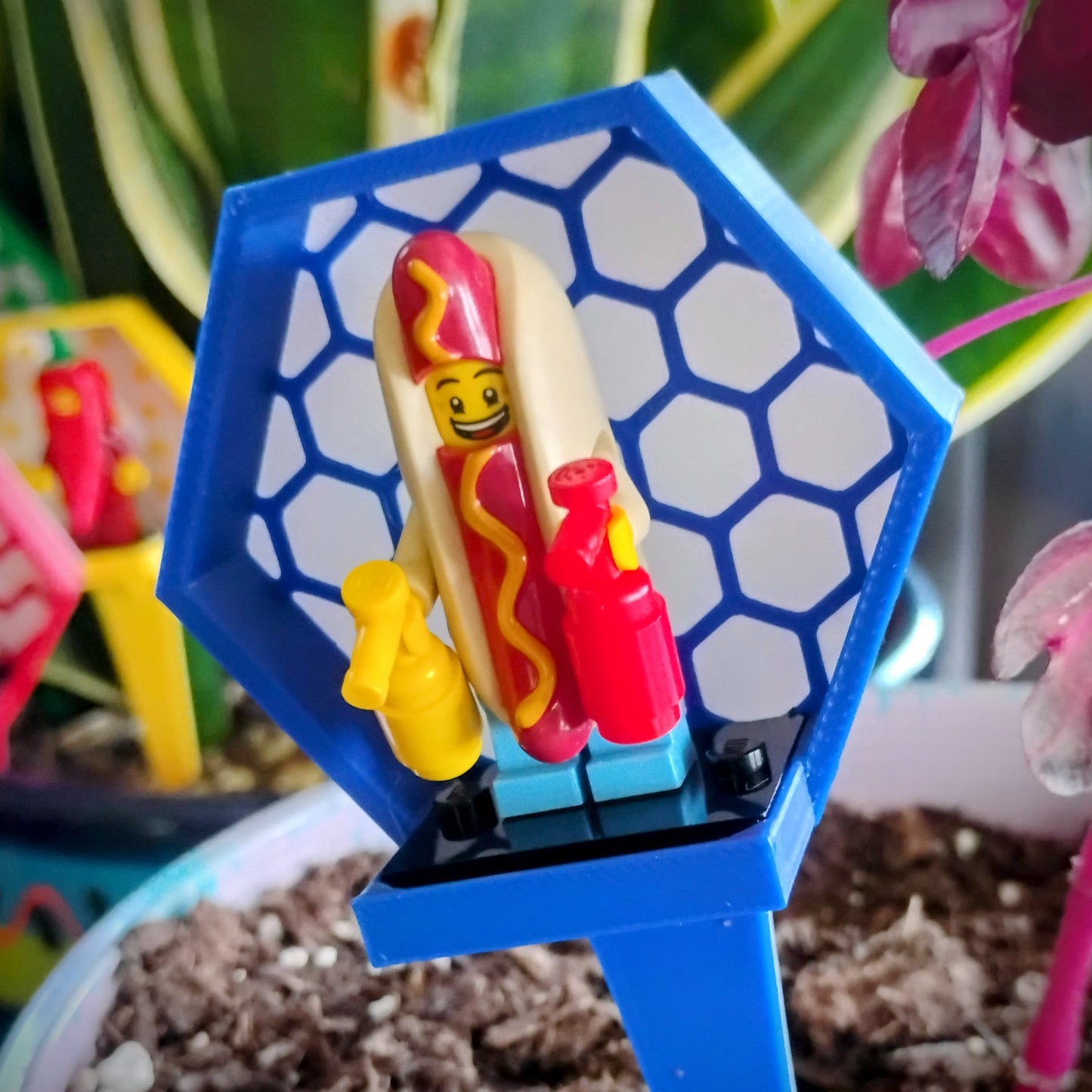 Close-up of a blue hexagon LEGO® minifigure plant stake holding a hot dog minifigure among houseplant leaves.