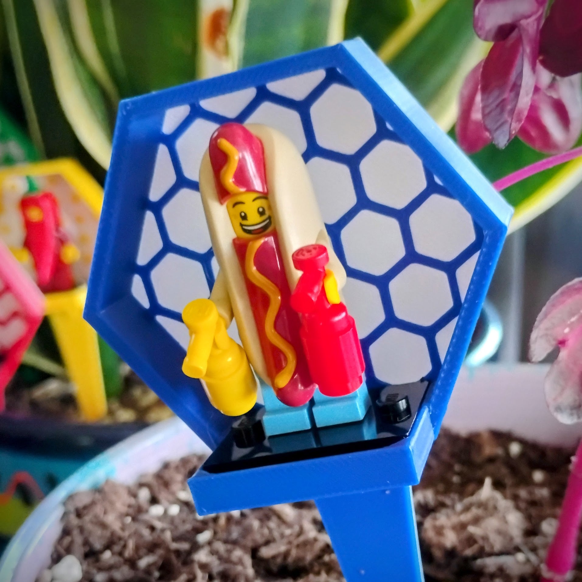 Close-up of a blue hexagon LEGO® minifigure plant stake holding a hot dog minifigure among houseplant leaves.