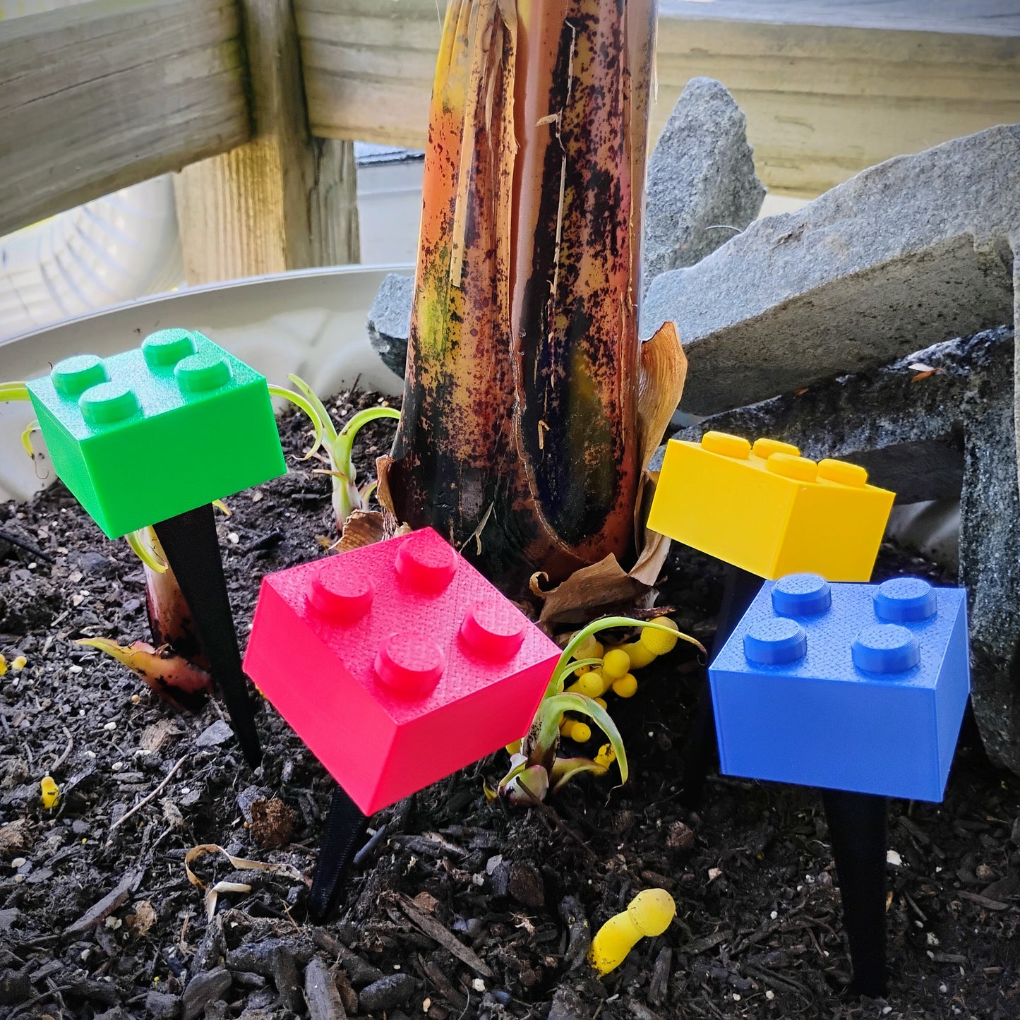 Colorful building brick plant stakes placed in the soil of an outdoor planter around a large plant.
