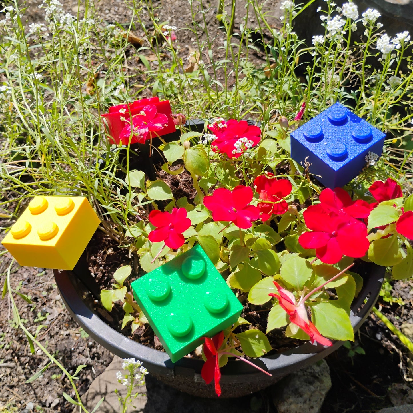 Red, blue, green and yellow building brick plant stakes displayed in a planter with red flowers.
