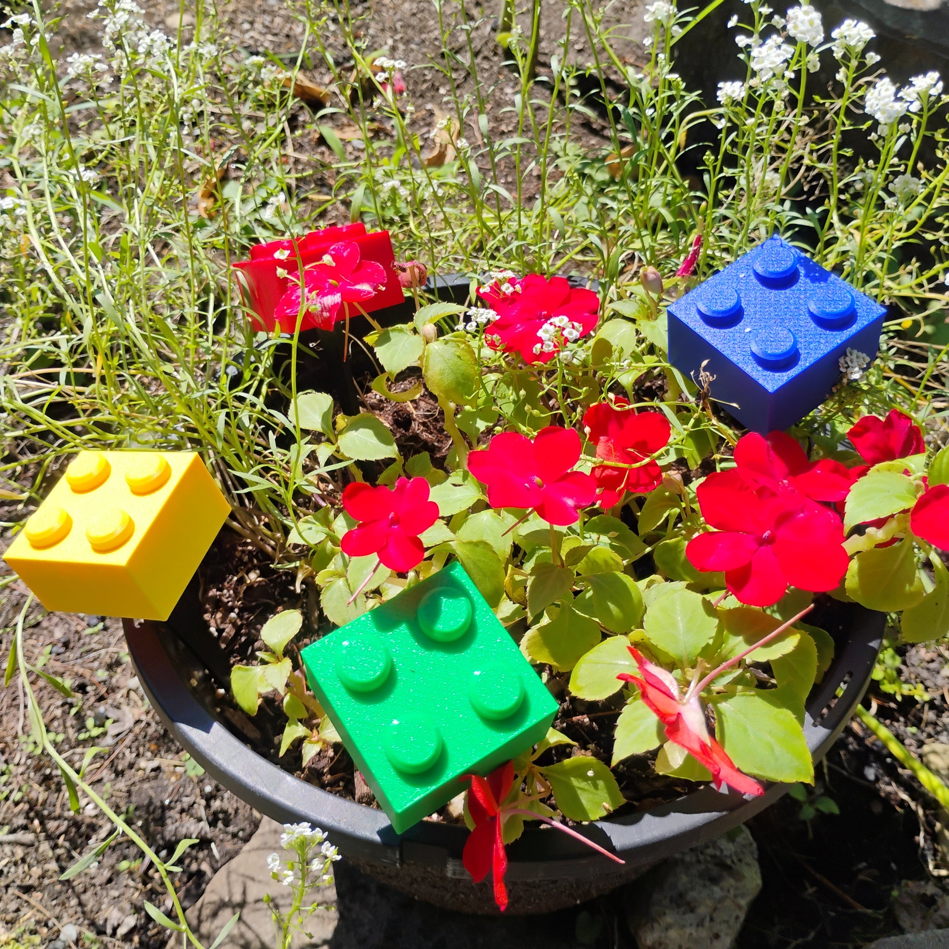 Red, blue, green and yellow building brick plant stakes displayed in a planter with red flowers.