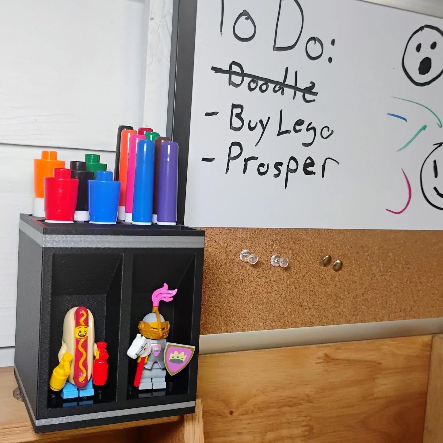 Black dry erase marker holder mounted near a classroom whiteboard and corkboard, organizing colorful markers while displaying two LEGO® minifigures.