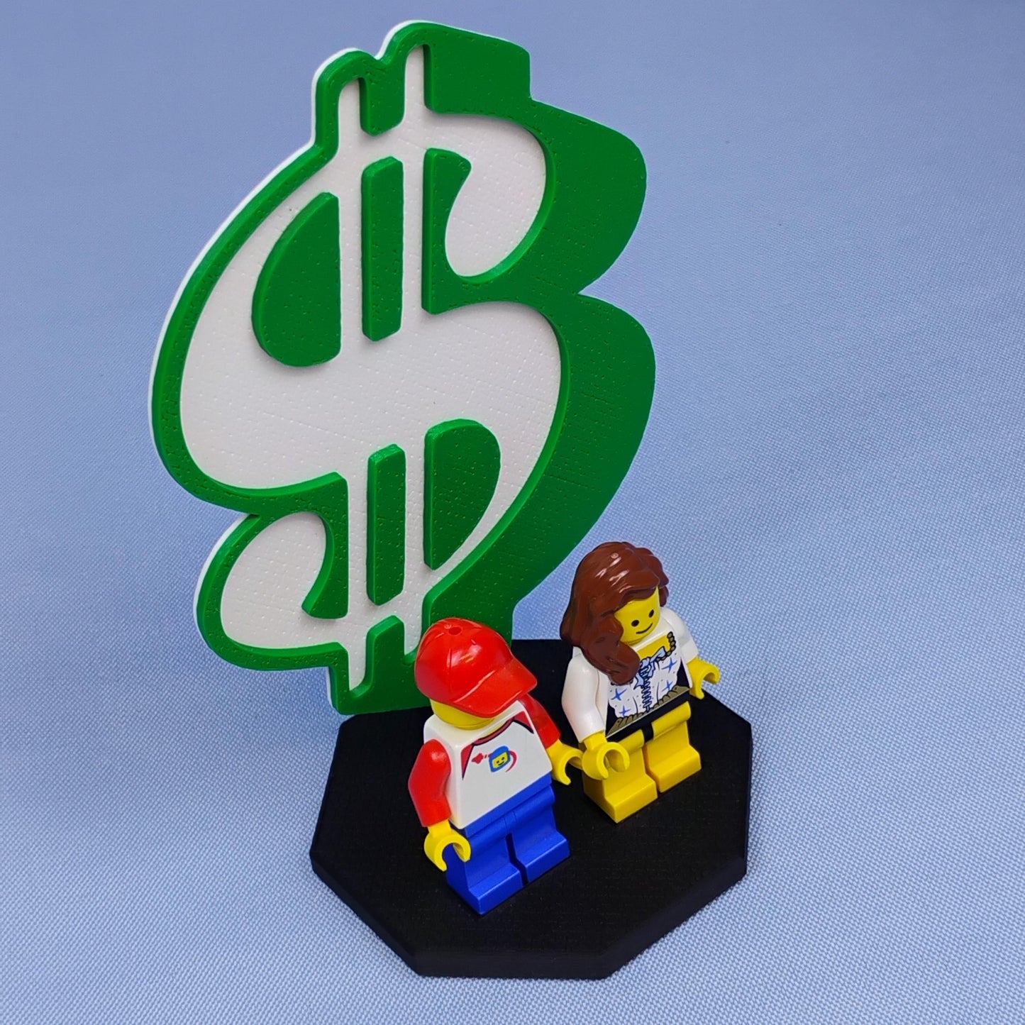 Dollar sign display stand for LEGO® minifigures shown against a blue studio background with green backdrop and black base.