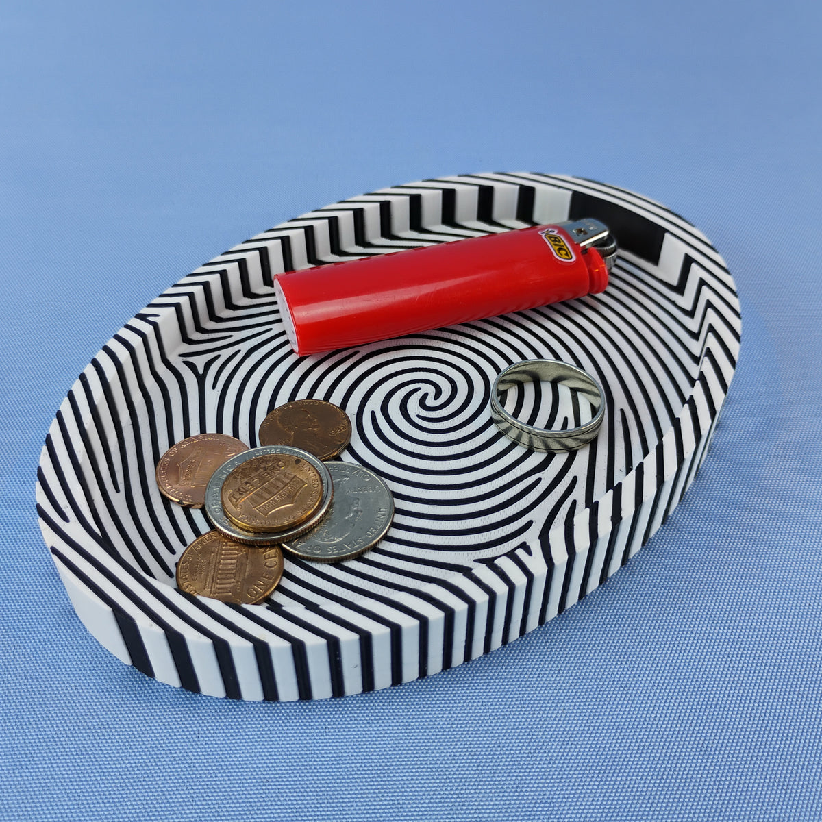 Black and white fingerprint catchall tray holding coins, a silver ring, and a red lighter on a blue background.