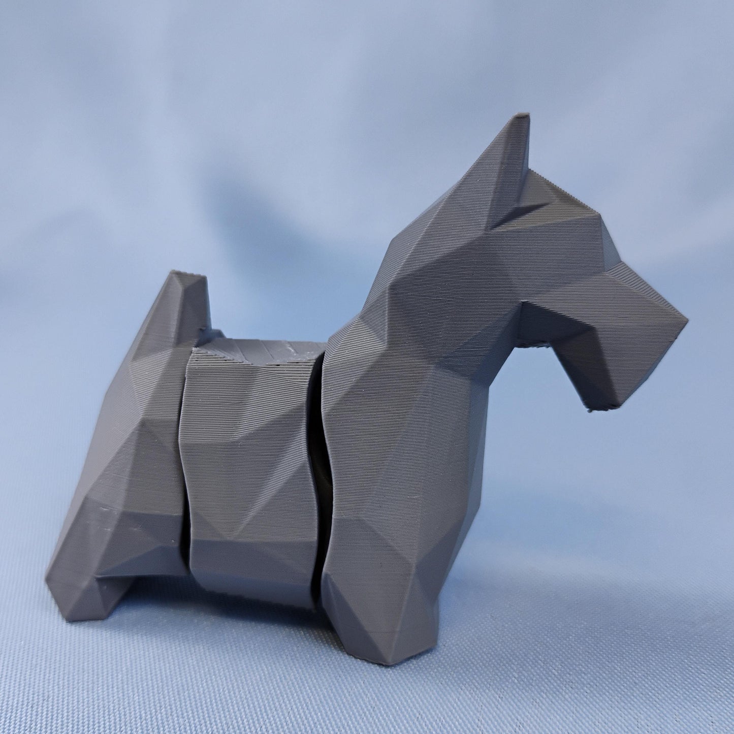 Flexi low-poly Scottish Terrier figurine in gray with faceted geometric details, shown on a blue background as desk or shelf decor.