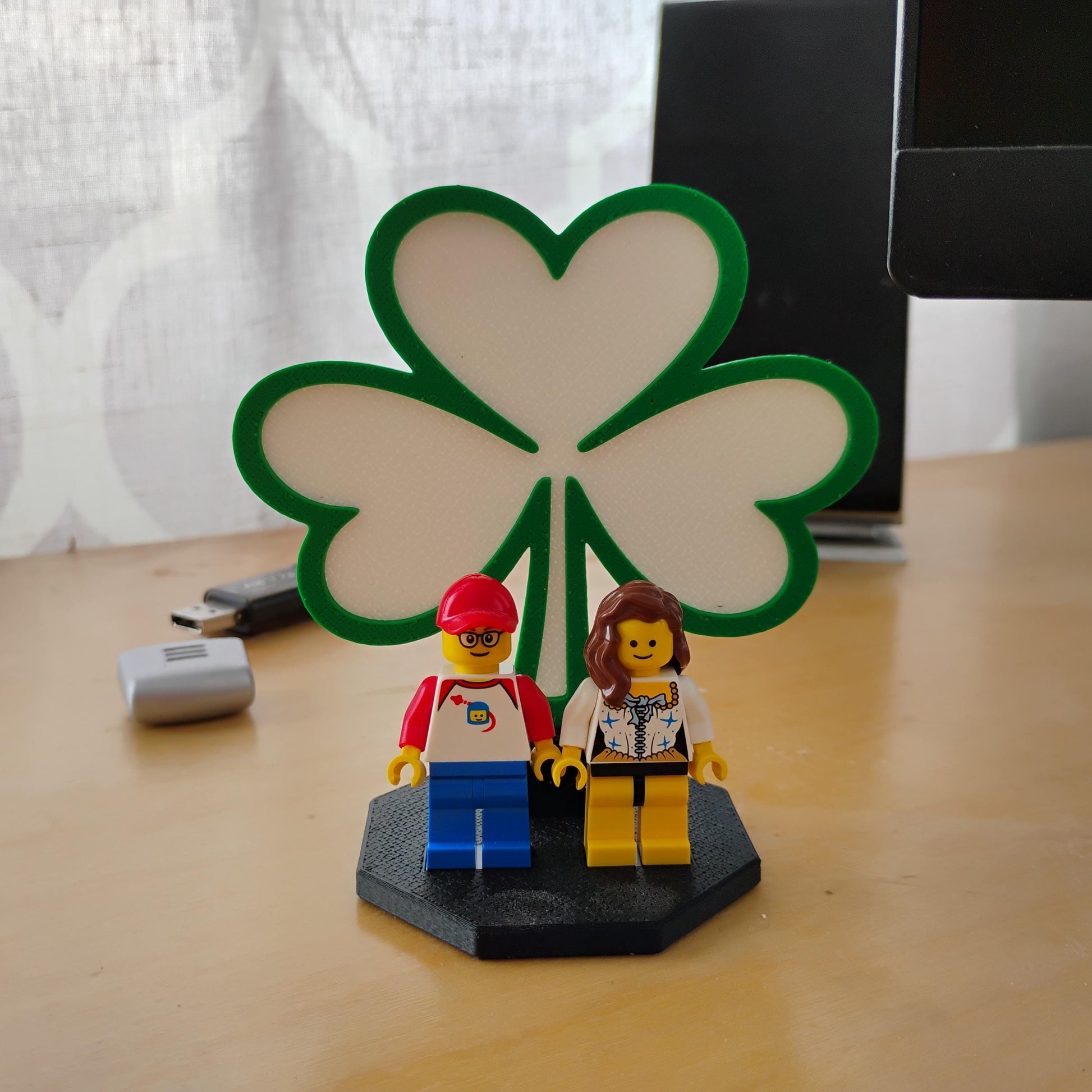 Green shamrock display stand for LEGO® minifigures shown on a desk with two figures and a black display base.