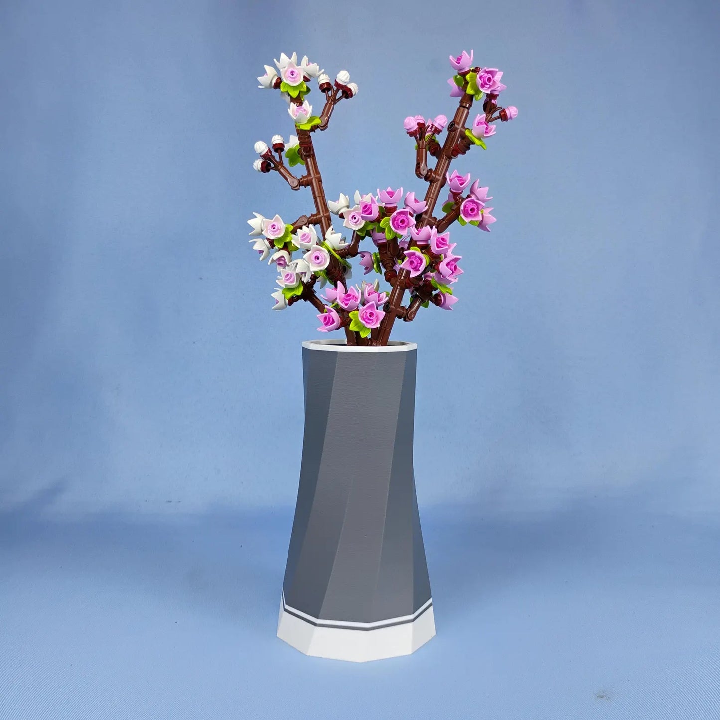 Grey faceted vase displaying pink LEGO® Botanical cherry blossom stems, shown against a soft blue background for desk or shelf decor.