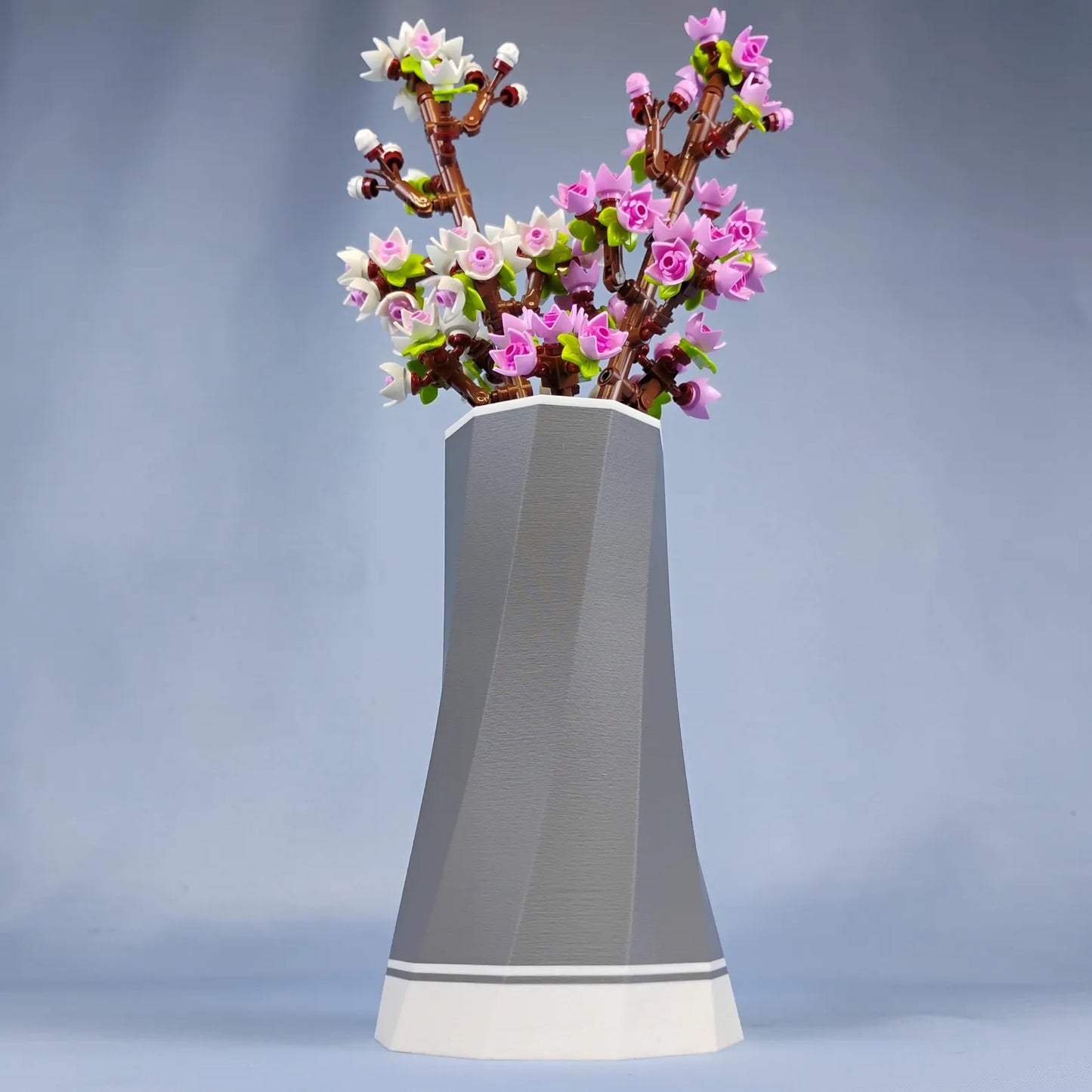 Grey faceted LEGO® Botanical vase with white accent base displaying pink cherry blossom stems against a blue studio background.