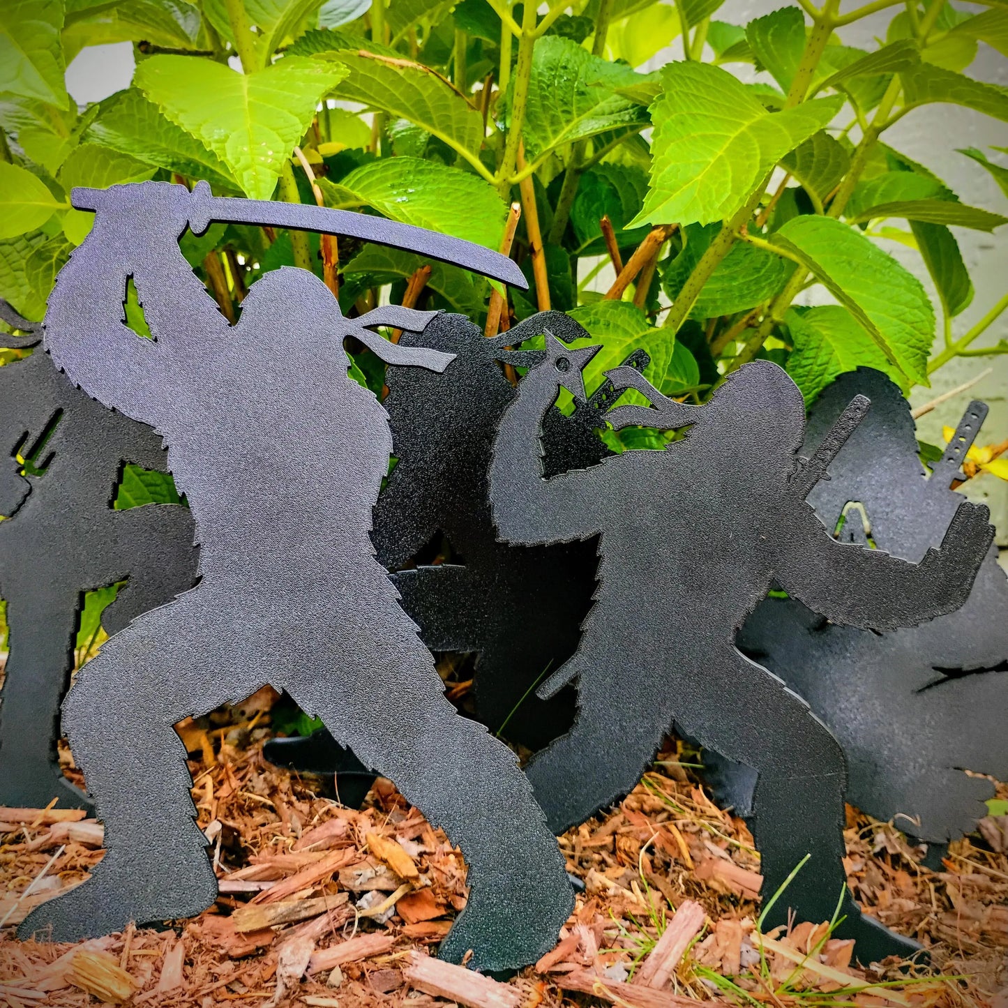 Complete set of five black Ninja Bigfoot Garden Spikes displayed together in a mulched garden bed among green plants.
