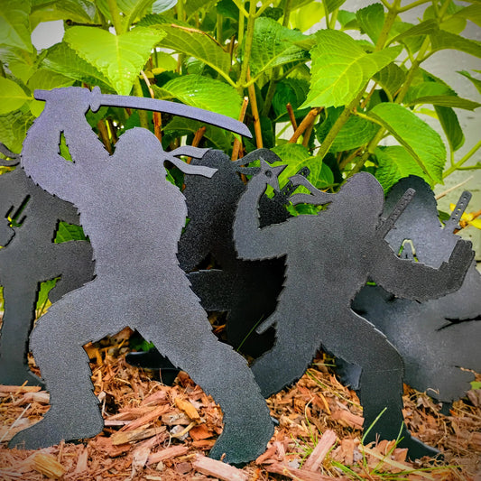 Complete set of five black Ninja Bigfoot Garden Spikes displayed together in a mulched garden bed among green plants.