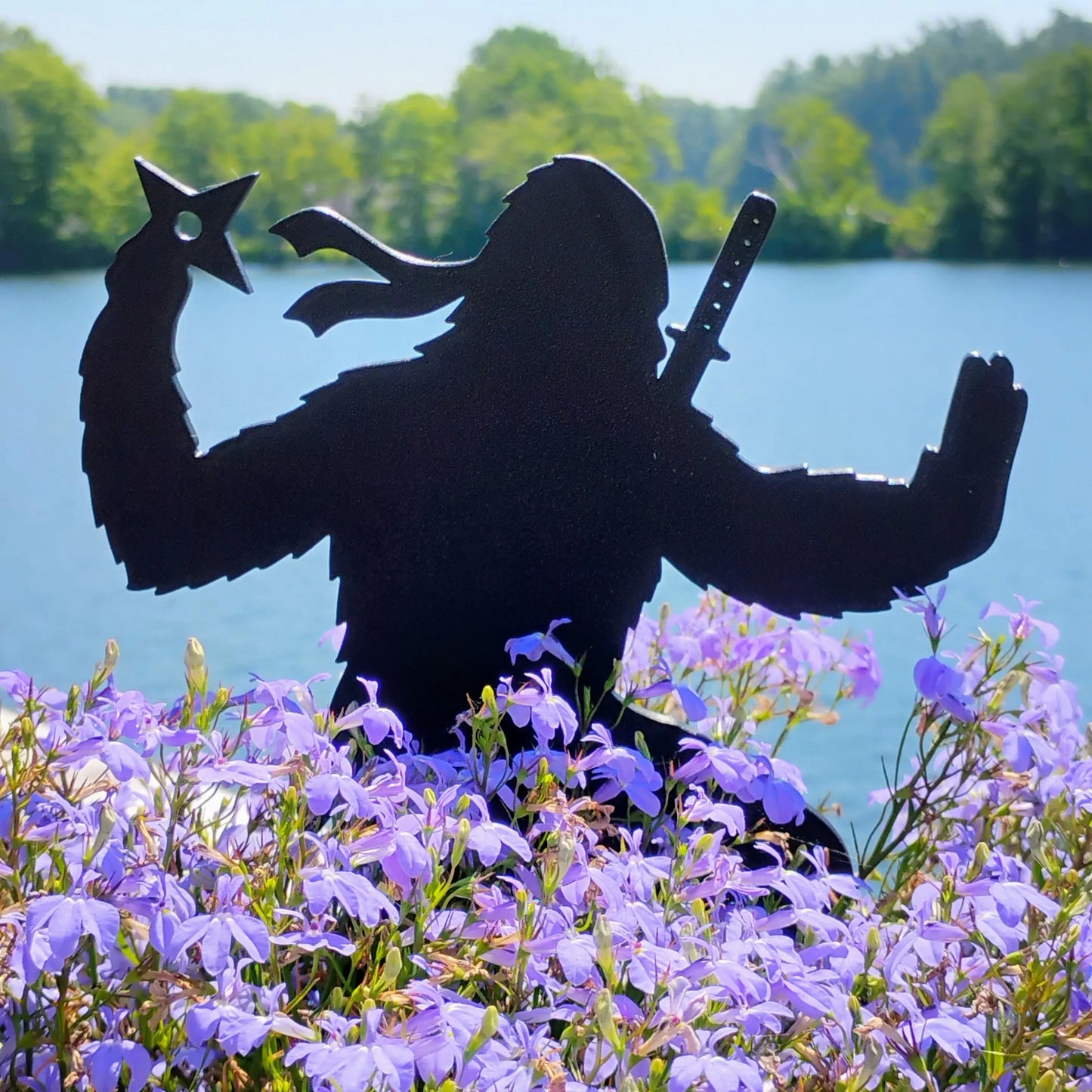 The Pointy One Ninja Bigfoot garden decoration holding throwing stars above purple flowers near a lake.