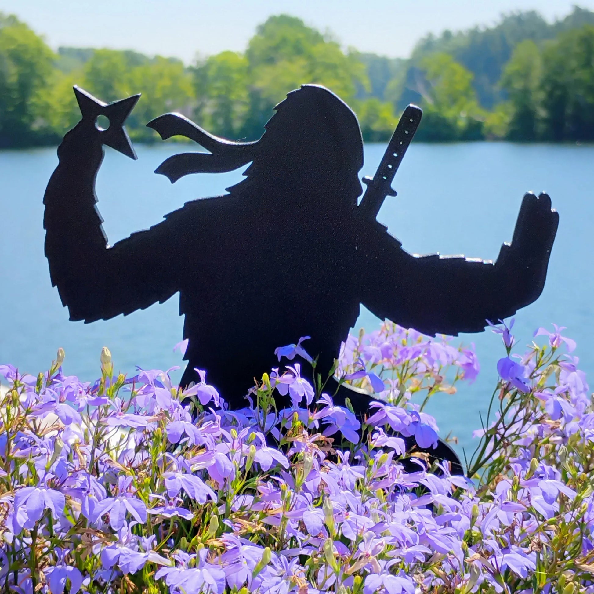 The Pointy One Ninja Bigfoot garden decoration holding throwing stars above purple flowers near a lake.