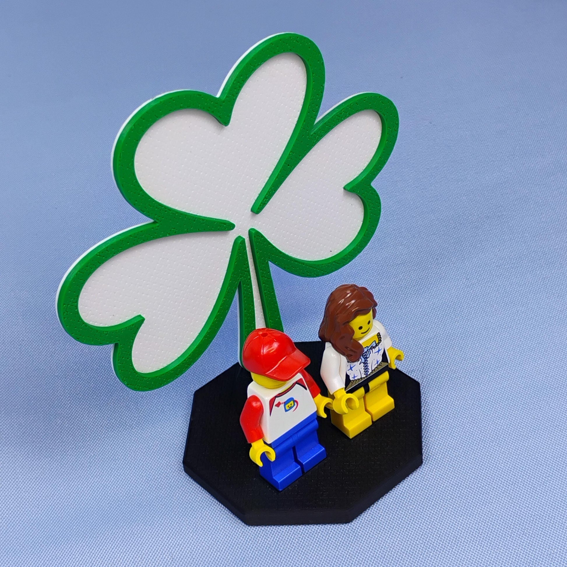 Shamrock display stand for LEGO® minifigures shown against a blue studio background with green clover backdrop and black base.