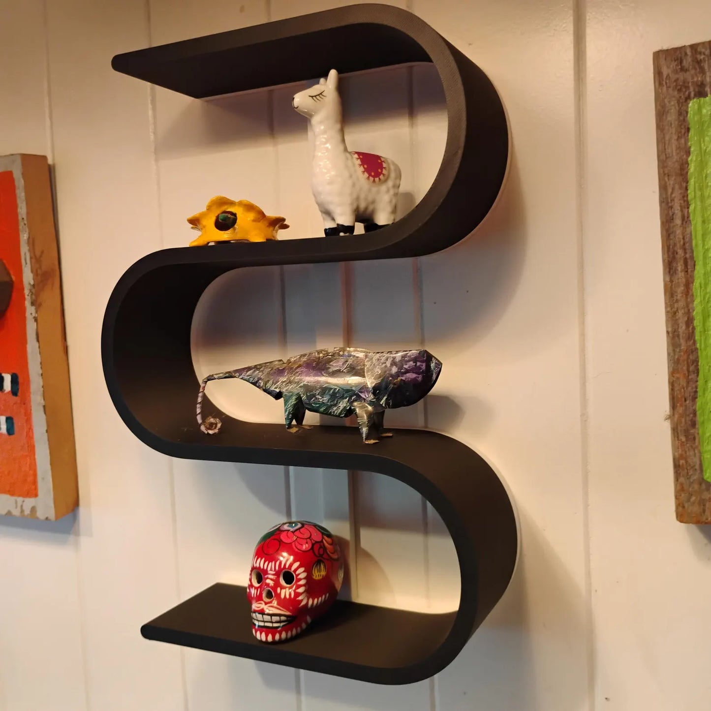 Black squiggly shelf mounted on a wall and styled with small oddities, animal figures, a miniature skull, and decorative collectibles across three curved levels.