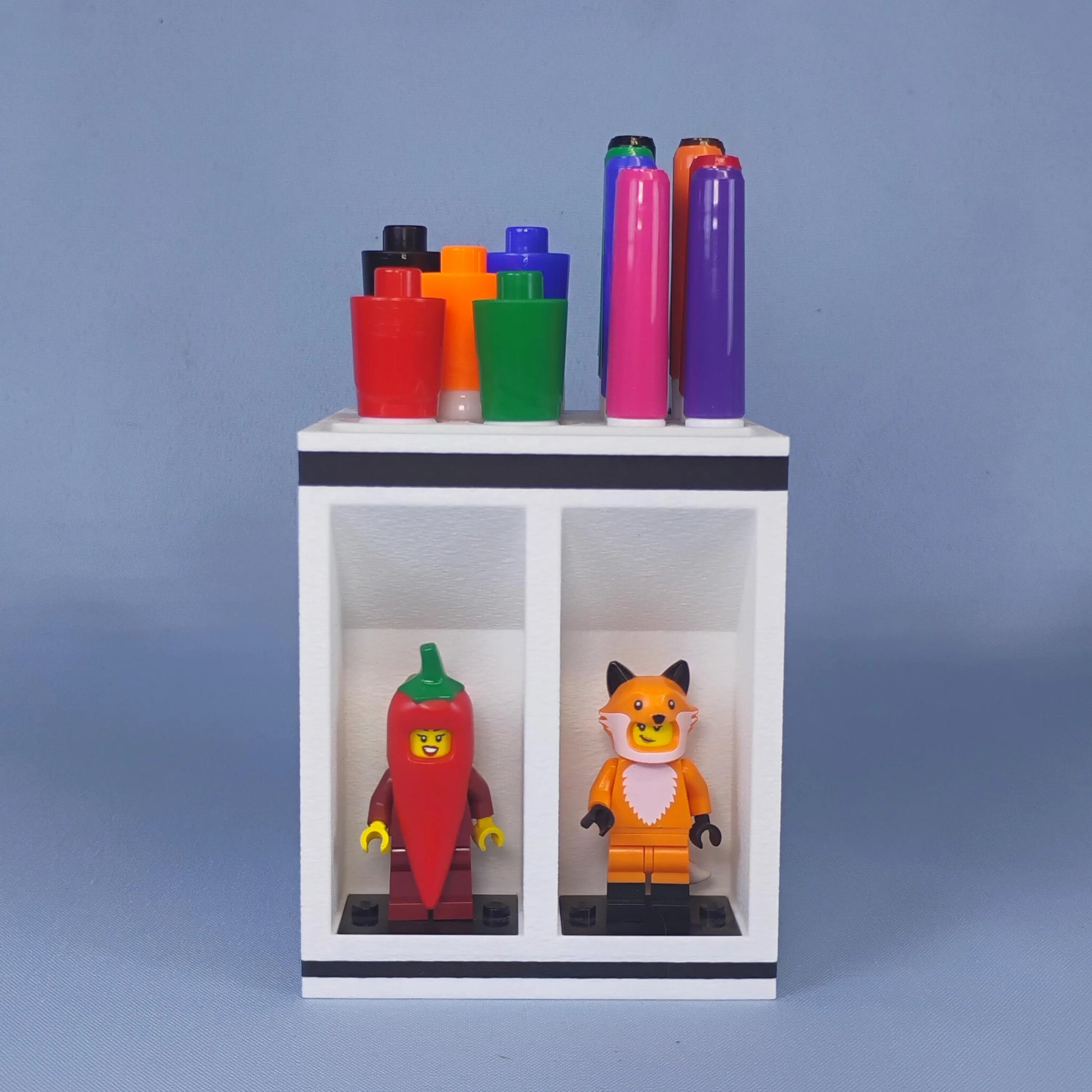 Front view of a white dry erase marker holder with large and fine-tip markers organized on top and two LEGO® minifigures displayed below.