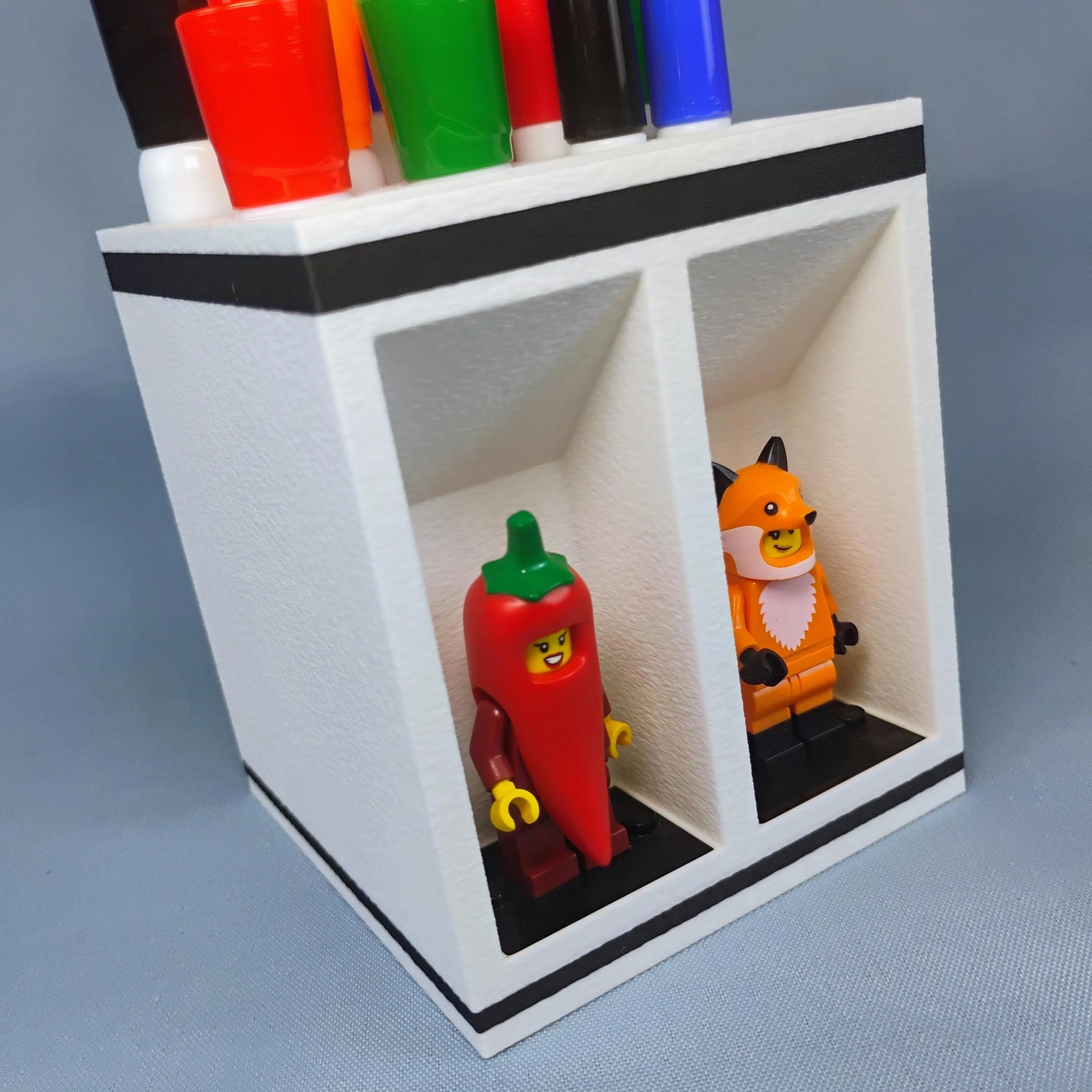 Close-up of a white dry erase marker holder with colorful markers stored on top and two LEGO® minifigures displayed in the open front bays.